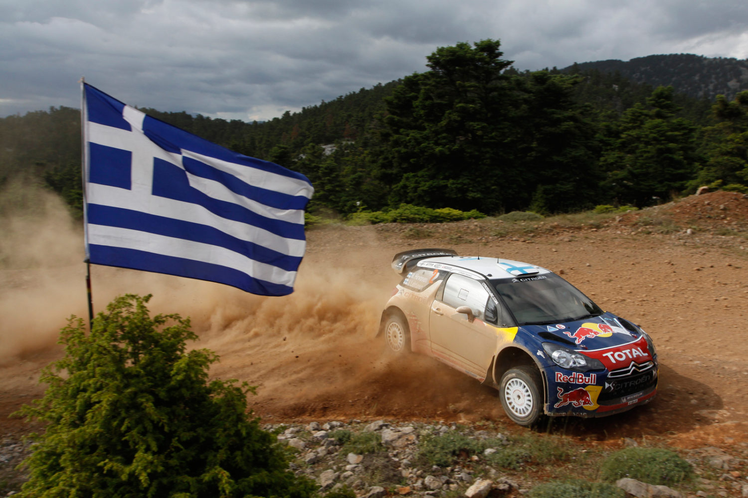 Voiture de rallye qui dérape dans le sable devant un drapeau de la Grèce